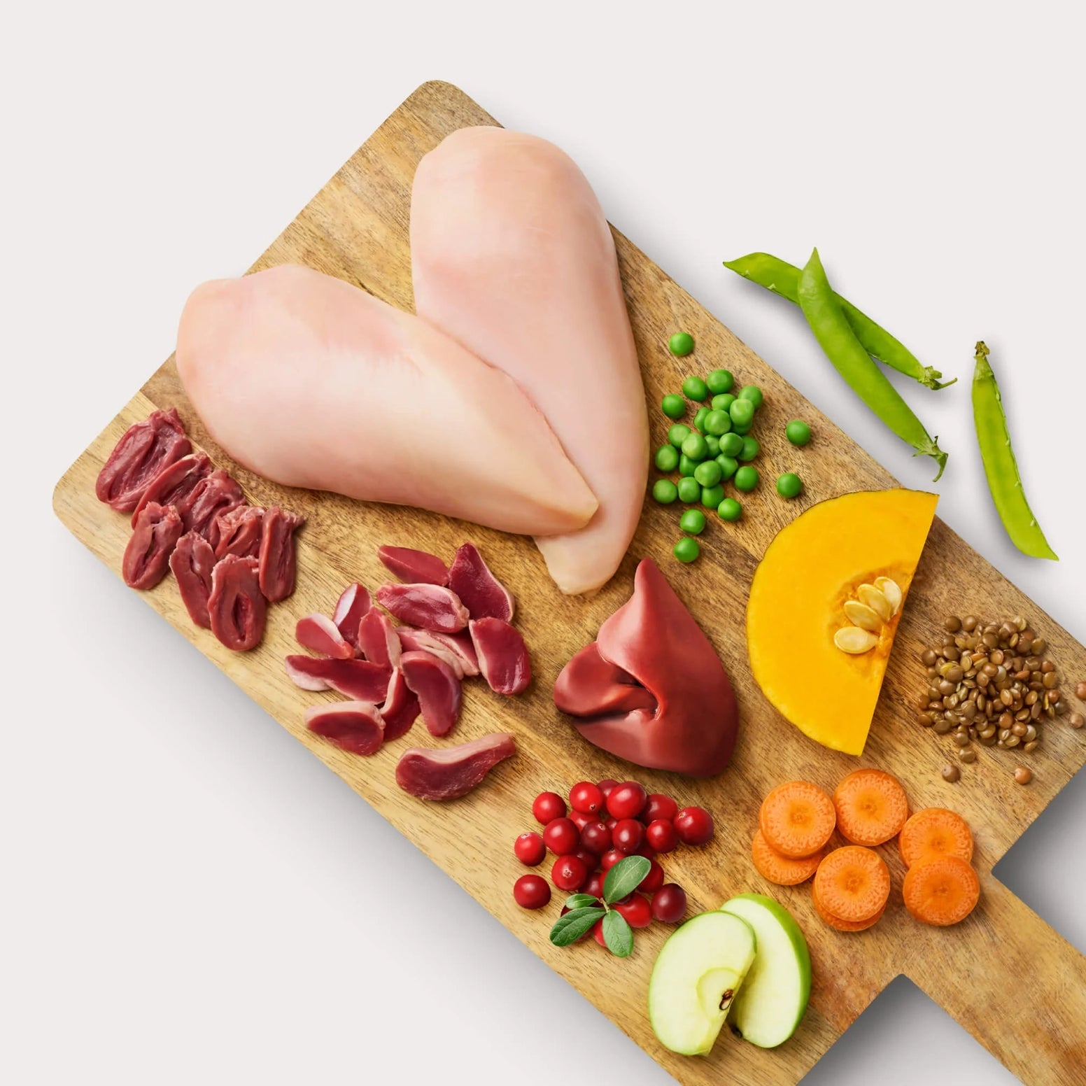 Raw chicken breast, organ meats, fresh vegetables, fruits, and seeds arranged on a wooden cutting board