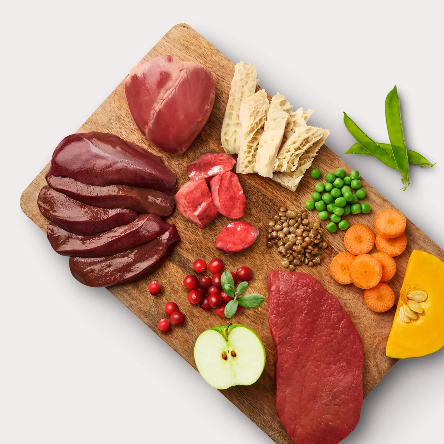 Assorted raw grass-fed beef cuts and fresh vegetables including peas, carrots, apple, and herbs on a wooden board