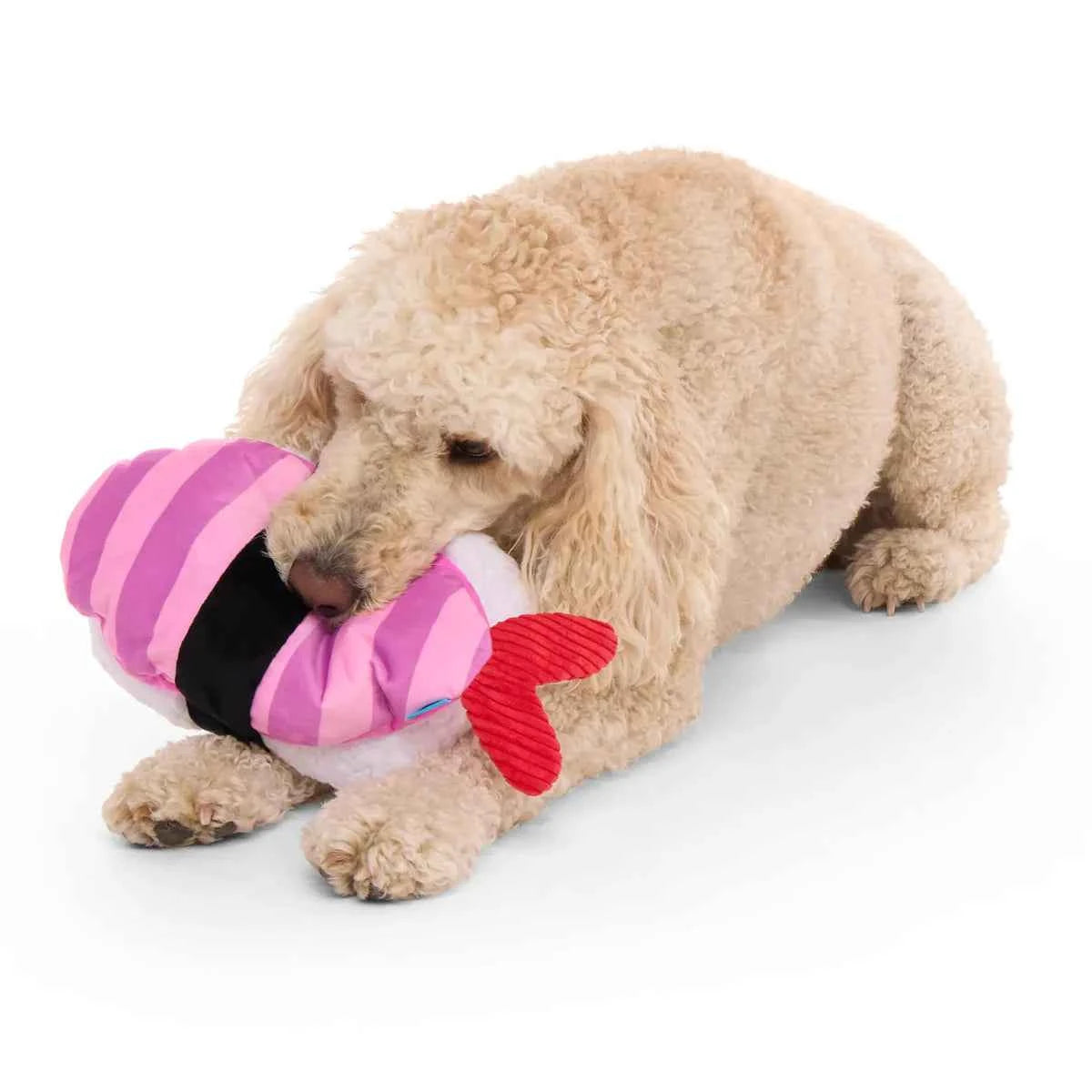 A large tan dog lying down and biting a pink and white Ebi sushi plush toy with a red tail.