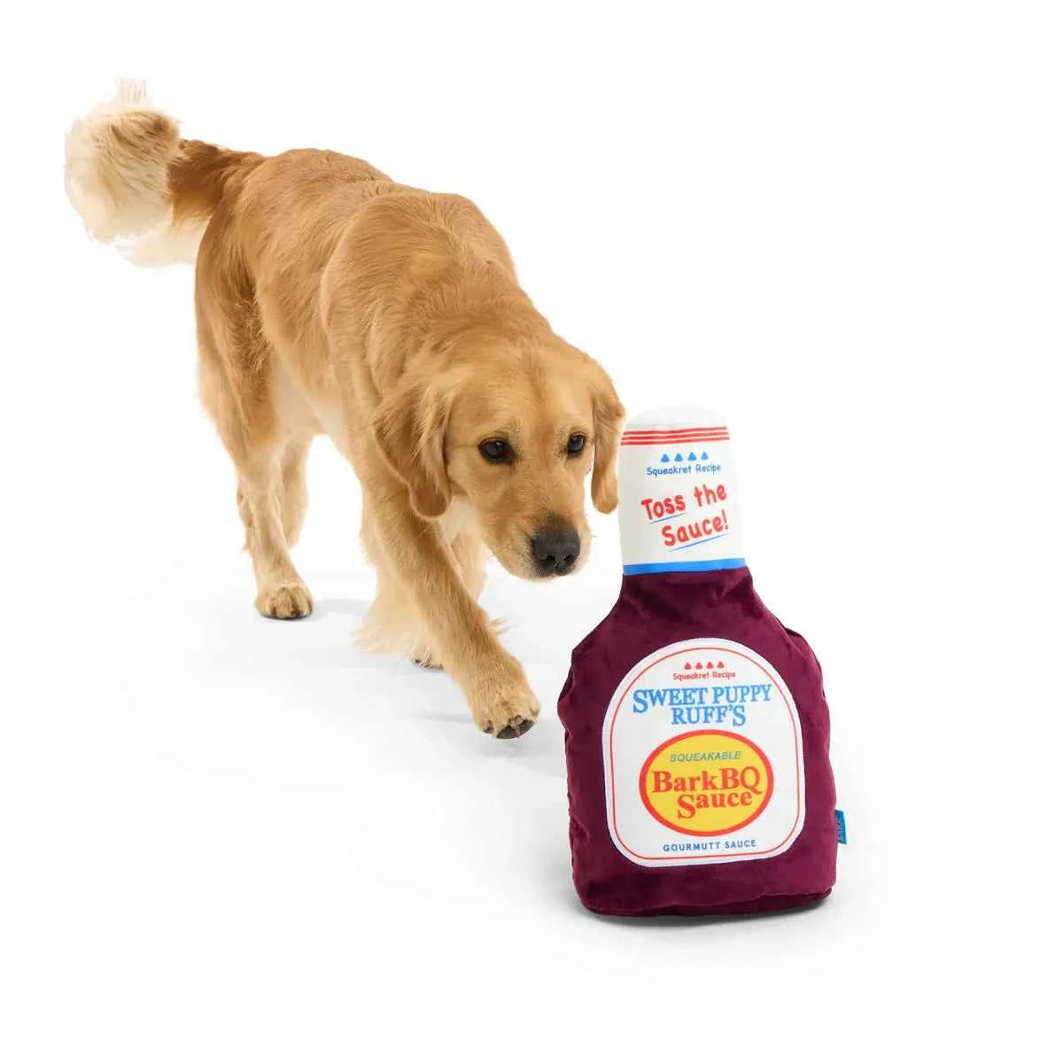 A golden retriever stands next to a large plush BarkBQ Sauce bottle dog toy.