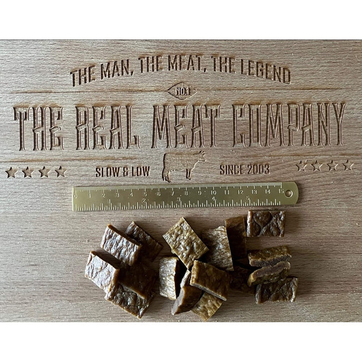 Square pieces of chicken and venison jerky dog treats on a wooden board next to a brass ruler.