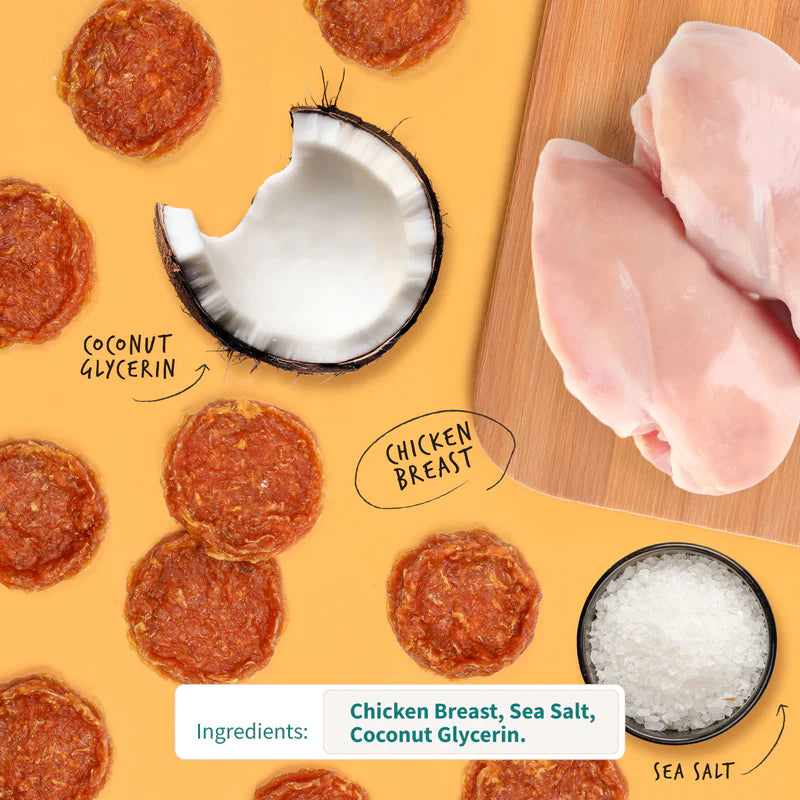 Round chicken-flavored dog treat chips surrounded by raw chicken breast, coconut half, and sea salt on soft orange background