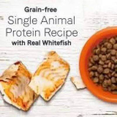 A 23.5 lb orange bag of CANIDAE Under The Sun Grain-Free Whitefish Recipe Dry Dog Food sits next to pieces of cooked whitefish on a white wooden background, with text highlighting its grain-free, single animal protein recipe.